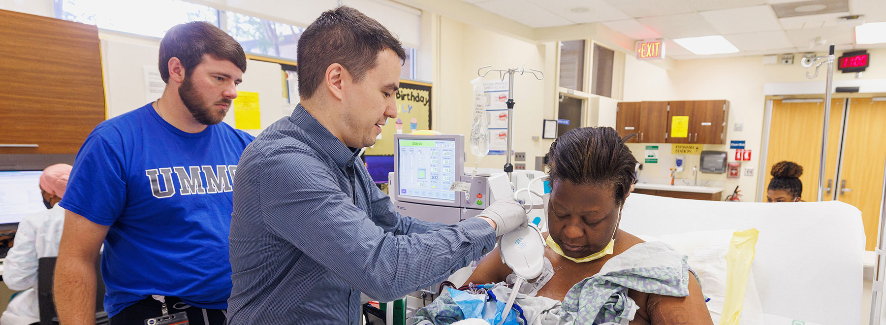 Healthcare professional adjusts a medical device connected to a patient seated in a hospital treatment room while another person observes nearby. Medical equipment, monitors, and clinical workstations are visible in the background.